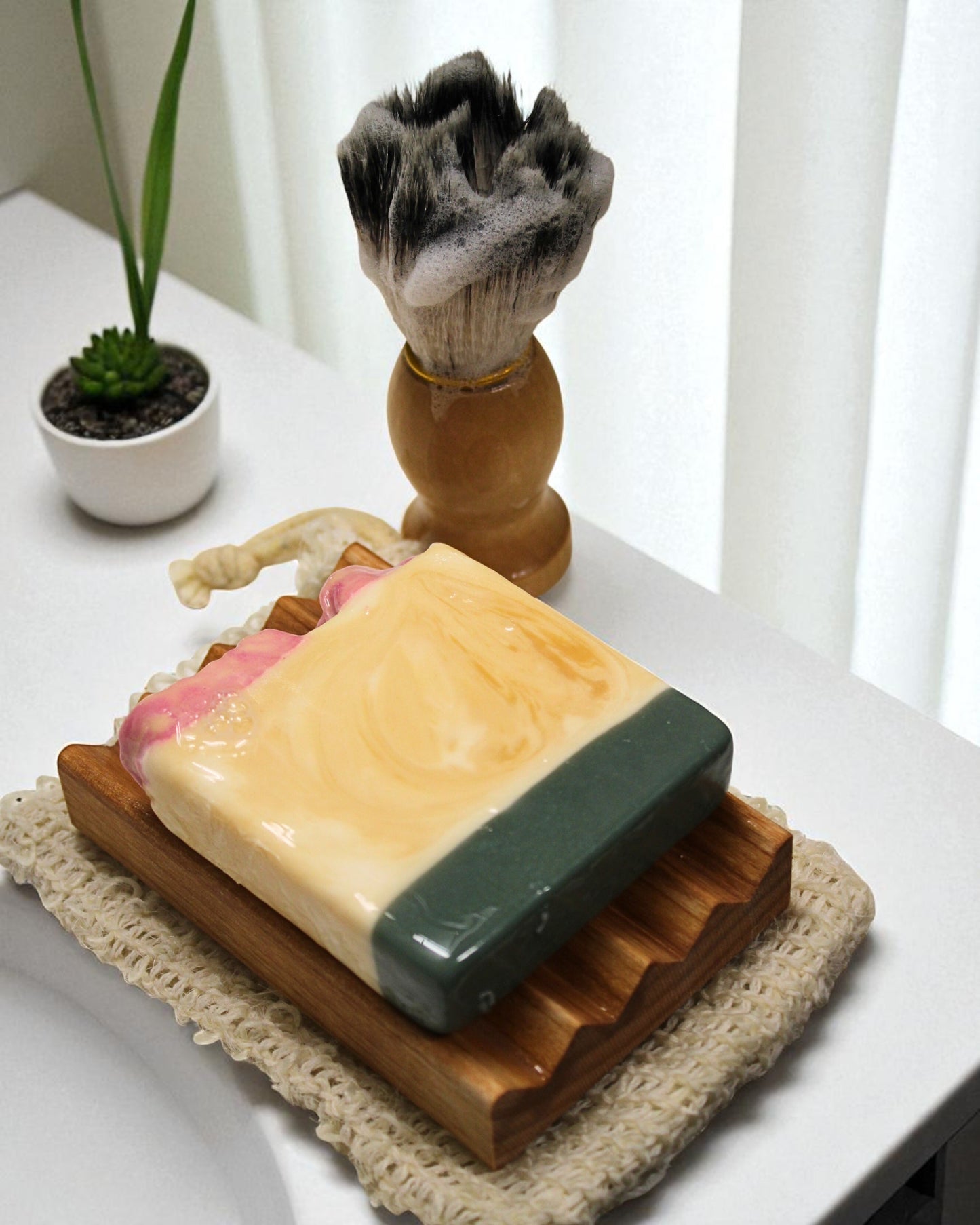 Bar of soap on a wooden soap dish with a shaving brush in the background, on a white surface.